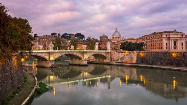 The Rome skyline reflects on the calm Tiber River at dusk with historic buildings and a bridge, HD Desktop Wallpaper