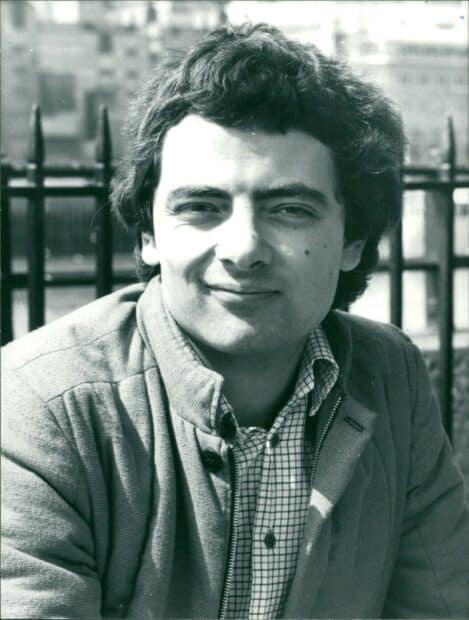 A young Rowan Atkinson smiles softly wearing a jacket outdoors with a fence in the background, HD Mobile Wallpaper