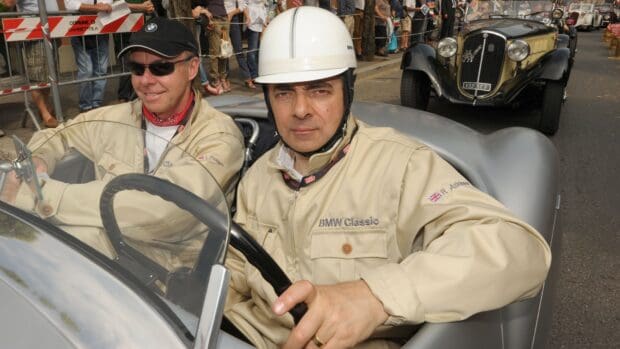 Rowan Atkinson is driving a vintage silver car wearing a white helmet and beige jacket, HD Desktop Wallpaper