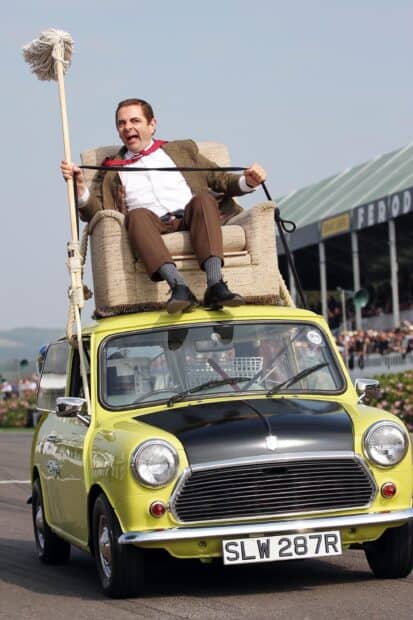 Rowan Atkinson is sitting on an armchair mounted on a yellow classic car holding a mop, 2K Mobile Wallpaper