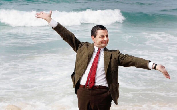 Rowan Atkinson posing by the sea with arms outstretched in a brown suit and red tie, 2K Desktop Wallpaper