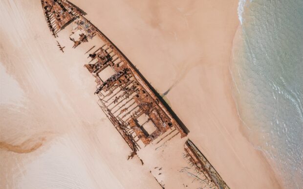 A rusty shipwreck lies partially buried on the sandy shore of Fraser Island, showing corroded metal and weathered wood, 2K Desktop Wallpaper