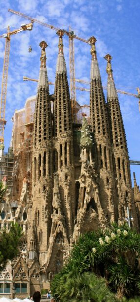 The Sagrada Familia towers rise majestically under a blue sky among green foliage, 2K Mobile Wallpaper
