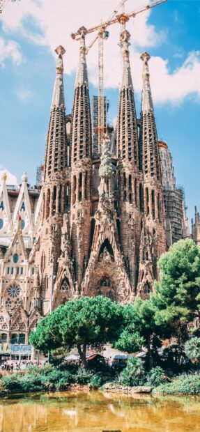 The Sagrada Familia shows its iconic towers surrounded by trees under a blue sky, 2K Mobile Wallpaper