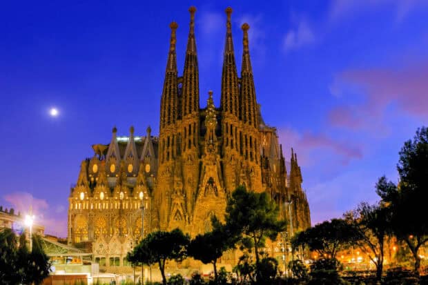 The Sagrada Familia shines brightly under a clear evening sky with surrounding trees, 2K Desktop Wallpaper