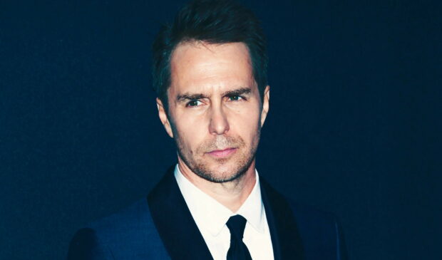 Sam Rockwell looks thoughtful in a dark suit and tie against a blue background, 5K Desktop Wallpaper