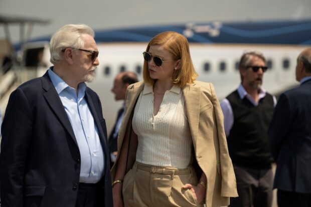 Sarah Snook is wearing a plaid suit and sunglasses while talking to a man near an airplane, HD Desktop Wallpaper