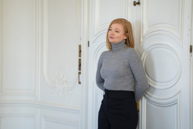 Sarah Snook wallpapers shows her leaning against a white ornate door wearing a gray turtleneck and black pants, HD Desktop Wallpaper