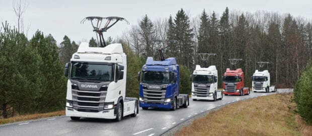 A convoy of Scania trucks drives along a road surrounded by trees under a cloudy sky, 2K Desktop Wallpaper
