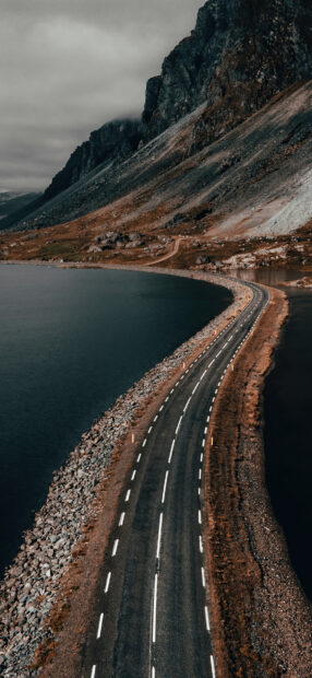 A winding coastal road runs alongside rocky shores beneath towering mountains in Norway, HD Phone Wallpaper