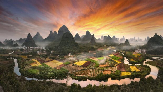 A peaceful village surrounded by green mountains under a vibrant sunset sky, HD Desktop Wallpaper