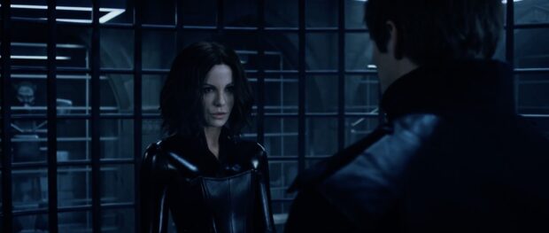 Selene from Underworld wears a black leather outfit standing inside a metal barred room with a man nearby, 2K Desktop Wallpaper