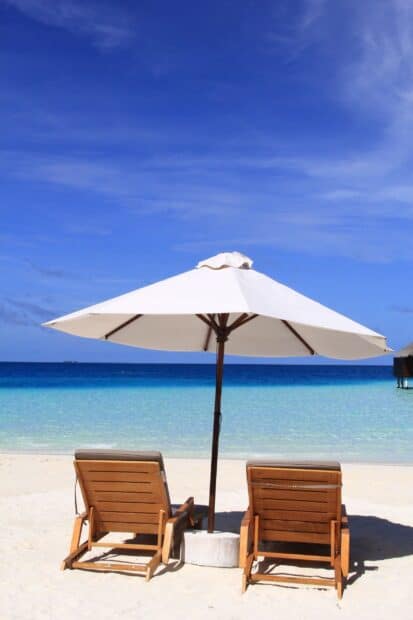A beach umbrella stands between two wooden chairs on the white sand shore, 2K Mobile Wallpaper