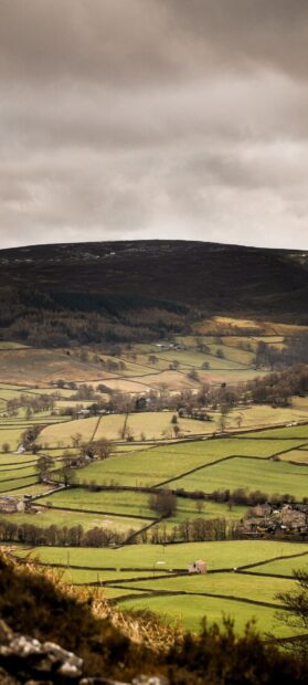 A peaceful village surrounded by patchwork green fields under a cloudy sky with hills, HD Phone Wallpaper