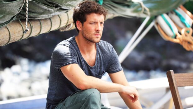 A serious man from Death In Paradise Tv Series sitting near a wooden structure outdoors, HD Desktop Wallpaper