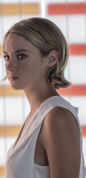 A serious woman wears a white top standing in front of a patterned window in Allegiant Wallpapers, 2K Phone Wallpaper