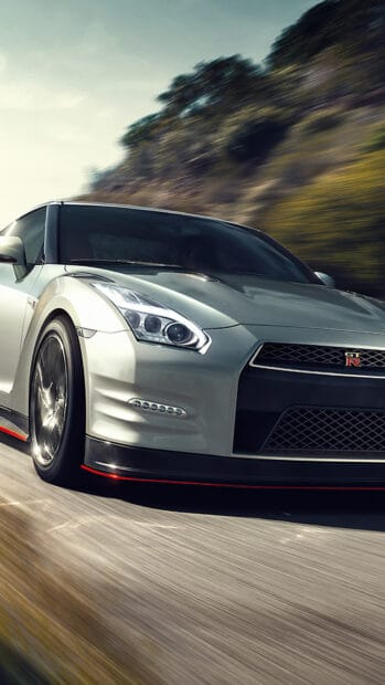 A silver Nissan GT R is driving fast along a scenic road with blurred trees in the background, 4K Phone Wallpaper