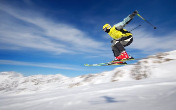 A skier wearing yellow gear is jumping high on a snowy mountain slope during a clear day, 2K Desktop Wallpaper