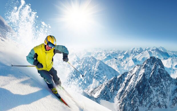 A skier wearing yellow jacket descends a snowy mountain slope with snowy peaks in the background, HD Desktop Wallpaper