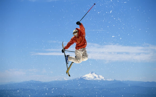 A skier is performing a jump trick in midair while snow is falling around and mountains are visible, HD Desktop Wallpaper
