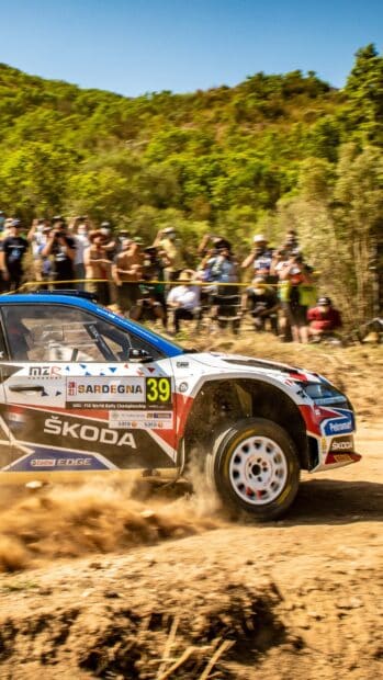 A Skoda racing car kicks up dust while navigating a rally raid course with spectators in the background, 2K Mobile Wallpaper