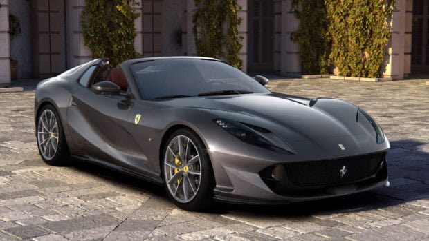 A sleek gray Ferrari 812 Gts convertible is parked on a stone driveway with greenery in the background