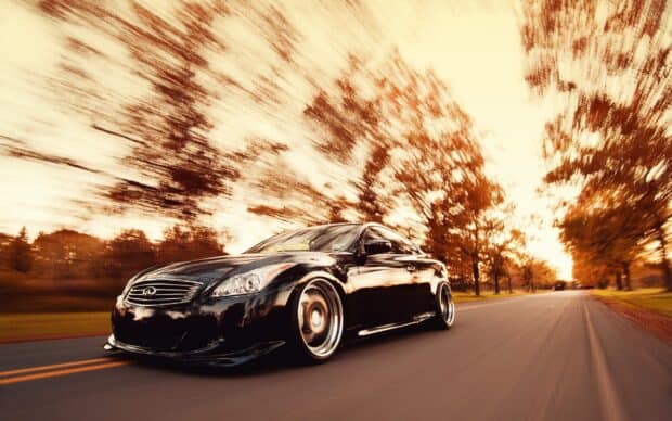 A black Infiniti G37 car is driving quickly down a tree lined road with autumn foliage blurred in the background, 2K Desktop Wallpaper
