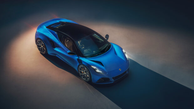 A sleek blue Lotus Emira is parked with its headlights on casting a long shadow, 2K Desktop Wallpaper