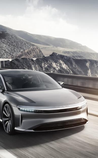 A sleek Lucid Motors car driving along a coastal highway with mountains in the background, 2K Mobile Wallpaper