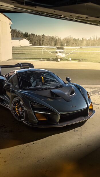 A sleek McLaren Senna sports car is parked inside a garage with an airplane visible outside, 4K Phone Wallpaper