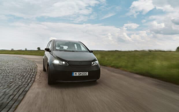 A black Sono Motors vehicle is driving quickly on a curved rural road under a partly cloudy sky, 2K Desktop Wallpaper
