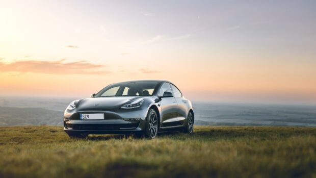 A sleek Tesla Model 3 is parked on a grassy field during sunset with an expansive landscape in the background, 2K Desktop Wallpaper