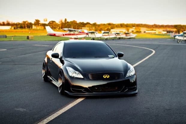 A sleek black Infiniti G37 is parked on an airstrip with small airplanes in the background, HD Desktop Wallpaper