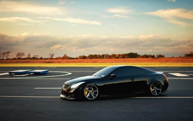 A sleek black Infiniti G37 is parked on an open asphalt area under a cloudy sky, 2K Desktop Wallpaper