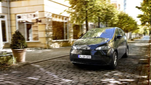 A sleek black Sono Motors vehicle drives on a cobblestone street surrounded by trees and buildings, 2K Desktop Wallpaper