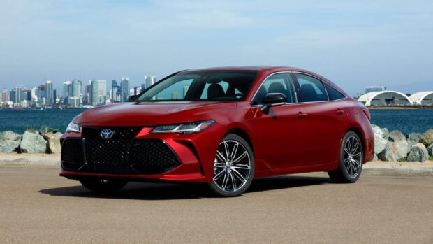 A sleek red Toyota Avalon is parked by the water with a cityscape in the background, HD Desktop Wallpaper