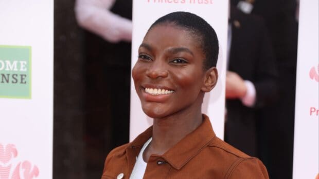Michaela Coel smiles warmly wearing a brown jacket at an outdoor event, 2K Desktop Wallpaper