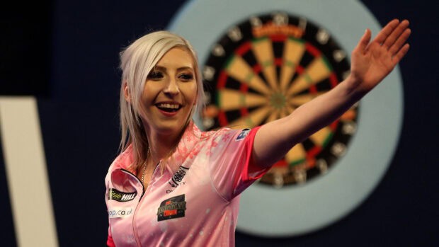 A female darts player wearing a pink shirt is smiling during a game in front of a dartboard, 4K Desktop Wallpaper