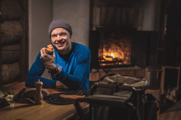 A man wearing a hat is enjoying a meal near a cozy fireplace in a wooden cabin featuring Iivo Niskanen wallpapers, 2K Desktop Wallpaper