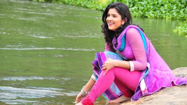 A smiling woman in traditional dress is sitting on a rock by the river in Bollywood Wallpapers, 4K Desktop Wallpaper