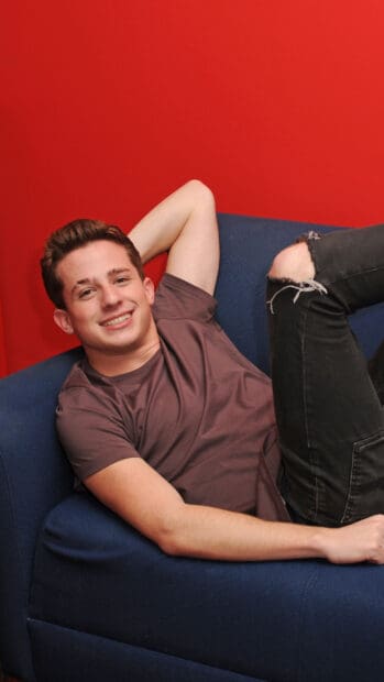 A young man smiles comfortably while resting on a blue couch against a red wall, 4K Mobile Wallpaper