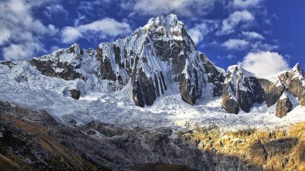 A snow covered mountain peak rises against a blue sky in Peru with rocky terrain below, 4K Desktop Wallpaper