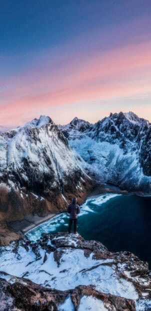 A person stands overlooking snowy glacier mountains and a blue sea under a pink sky, 2K Phone Wallpaper