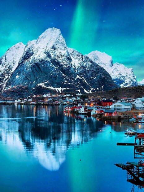 The snowy mountains reflect on calm water near a Norwegian town with northern lights in the sky, HD Mobile Wallpaper