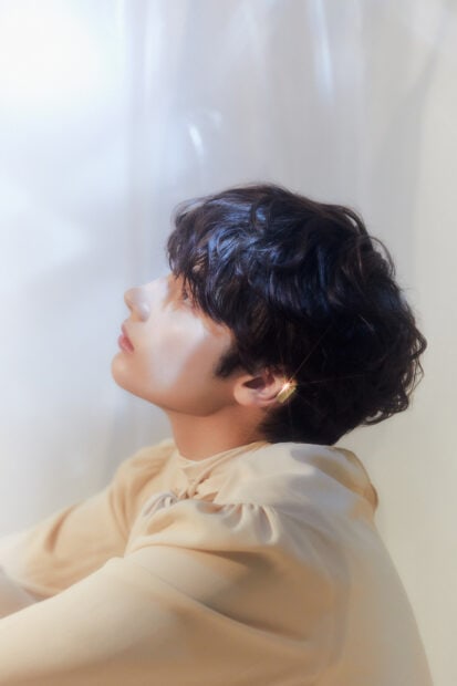 The image shows Huening Kai looking upward with soft lighting highlighting his curly hair and golden earring, HD Mobile Wallpaper
