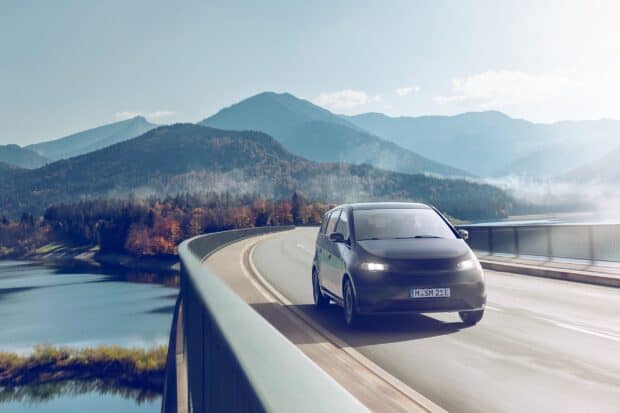 A Sono Motors vehicle drives on a curved mountain road with scenic forest and water views, 2K Desktop Wallpaper