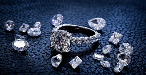 A sparkling diamond ring surrounded by various cut gems on a textured surface, HD Desktop Wallpaper