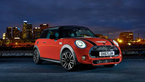 A red Mini Cooper with black stripes is parked on a dark street with city lights in the background, 4K Desktop Wallpaper