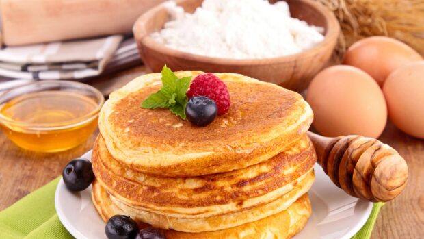 A stack of pancakes topped with fresh raspberries, blueberry, and mint leaves with honey and eggs nearby, 4K Desktop Wallpaper