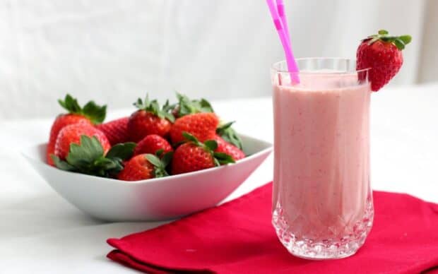 A glass of strawberry milkshake with a fresh strawberry garnish sits next to a bowl of ripe strawberries on a red napkin, 2K Desktop Wallpaper
